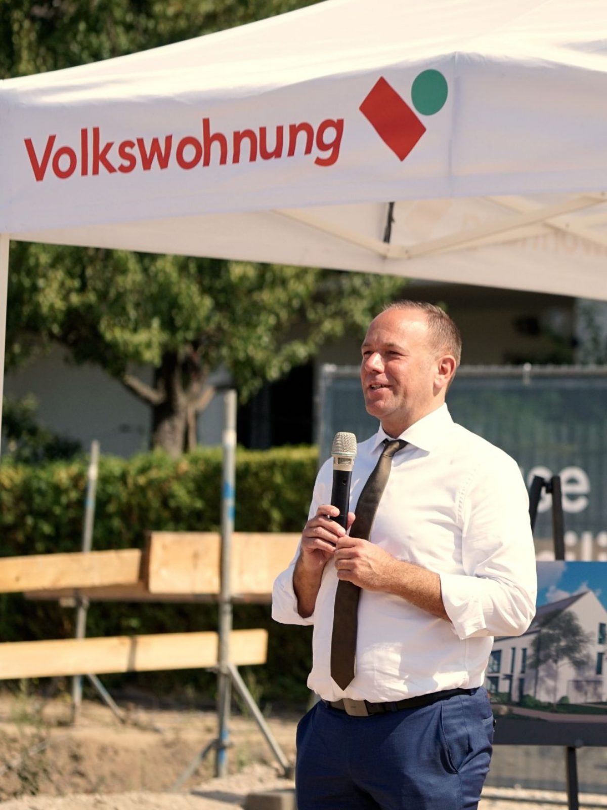 Ein Mann in weißem Hemd und Krawatte hält ein Mikrofon und spricht im Freien unter einem Zelt mit dem Logo der Volkswohnung. Im Hintergrund sind Bänke, Bäume und eine Baustelle zu sehen.