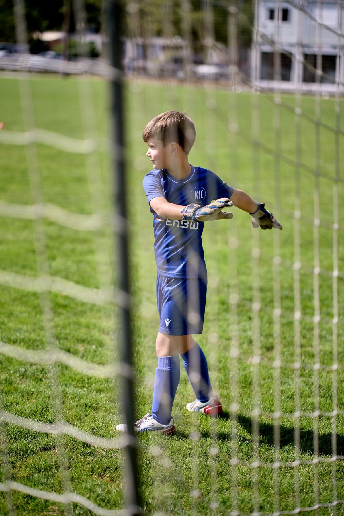 Ein Junge in einem blauen Fußballtrikot und Handschuhen steht in einem Tor auf einer Wiese und schaut mit ausgestreckten Armen zur Seite. Das Bild wird teilweise durch die Tornetze im Vordergrund eingerahmt.